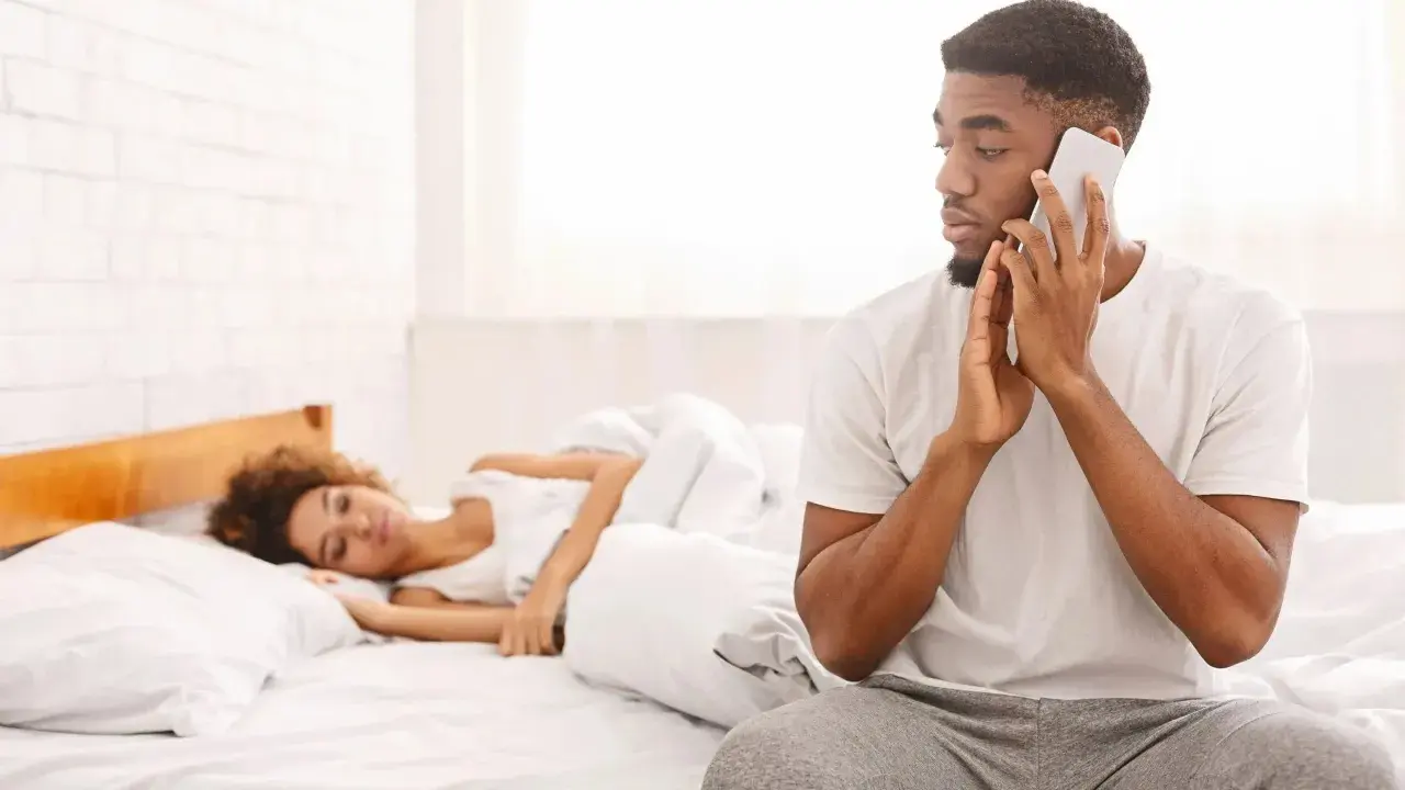 Un homme au téléphone, le regard fuyant, montre un comportement suspect alors qu'une femme dort paisiblement à côté de lui dans le lit.