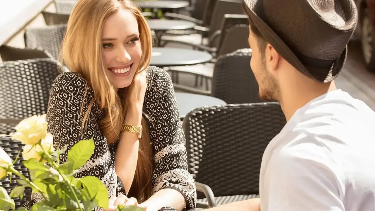 Un couple partage un moment tendre à une terrasse. Elle lui sourit, un bouquet de roses à la main, comme pour lui montrer comment le rendre accro.