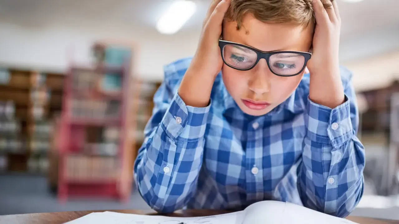 Garçon avec lunettes, mains sur la tête, visiblement en difficulté face à ses devoirs. Signes adolescent HPI, difficultés scolaires.