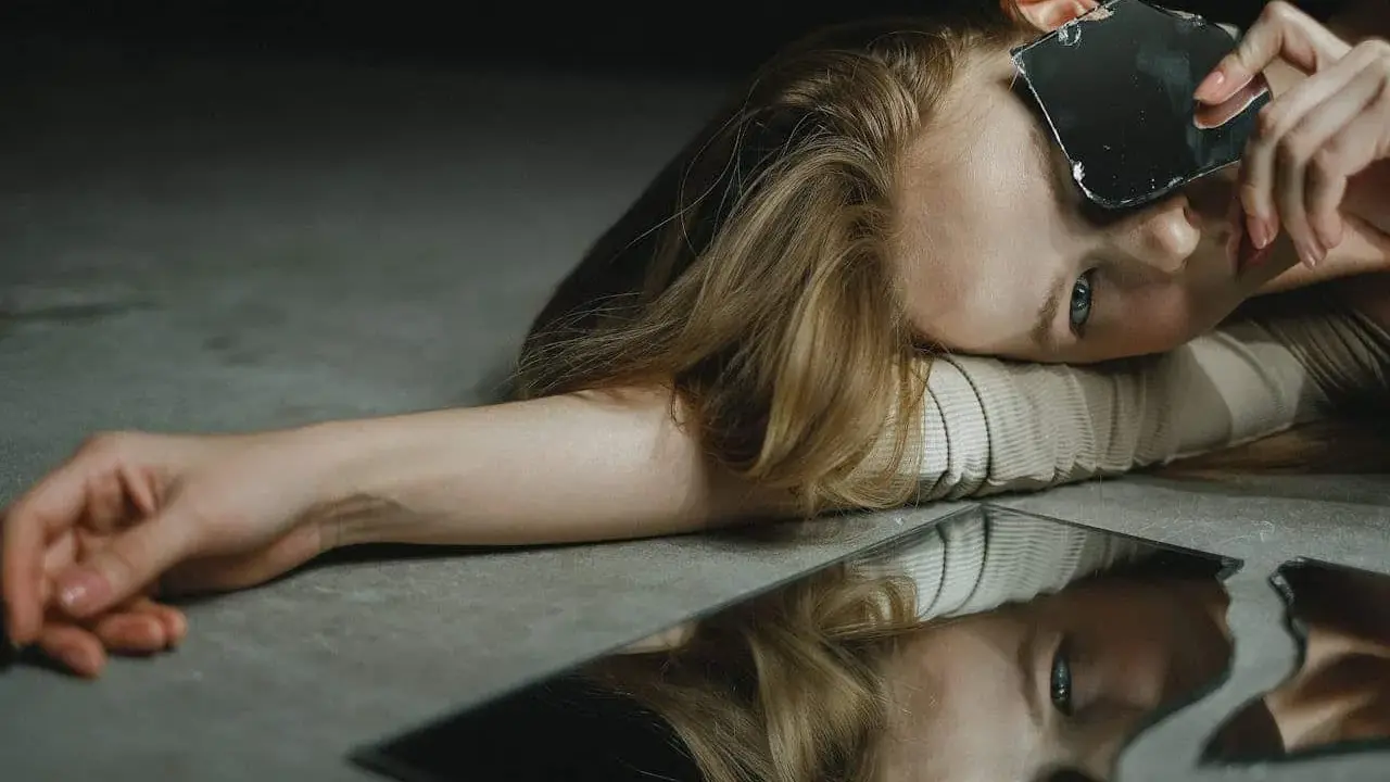 Une femme aux cheveux blonds, le visage tourné, tient un éclat de miroir. Son reflet brisé sur le sol évoque le narcissique et les femmes.