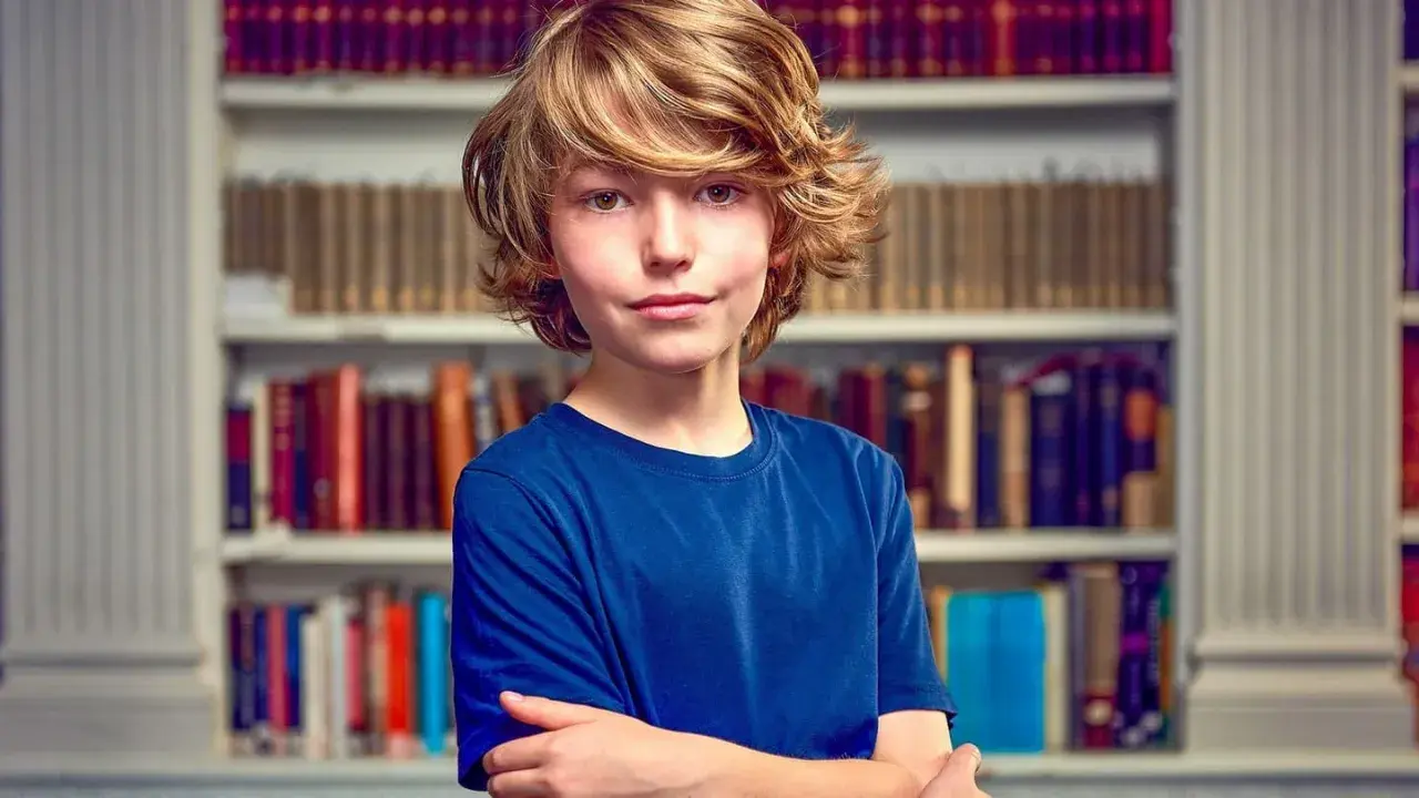 Garçon aux cheveux blonds, bras croisés devant une bibliothèque. Il semble prêt pour un test pour sauter une classe.
