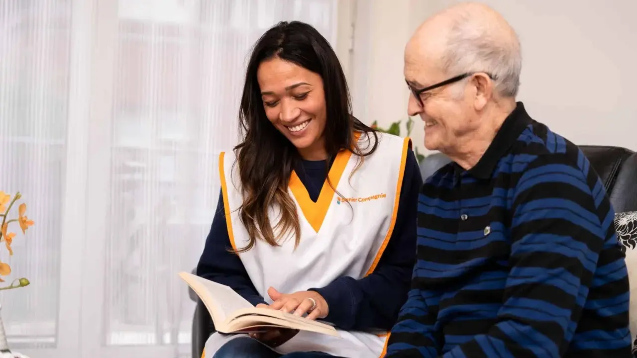 Une jeune femme souriante lit un livre à un homme âgé. Une scène qui illustre comment parler à une fille qu'on ne connaît pas : par le partage et l'écoute.