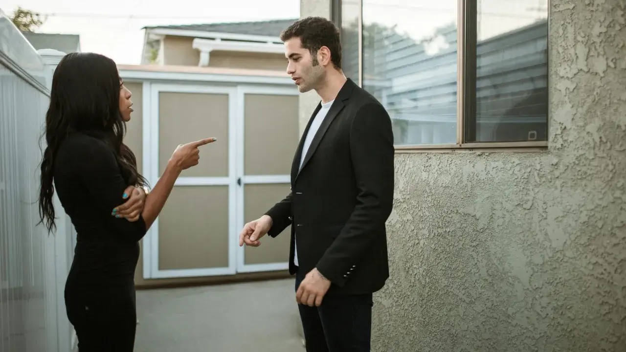 Une femme noire en robe noire pointe du doigt un homme en costume noir, lui reprochant quelque chose. Elle semble vouloir lui faire regretter quelqu'un.