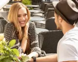 Un couple partage un moment tendre à une terrasse. Elle lui sourit, un bouquet de roses à la main, comme pour lui montrer comment le rendre accro.