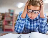 Garçon avec lunettes, mains sur la tête, visiblement en difficulté face à ses devoirs. Signes adolescent HPI, difficultés scolaires.