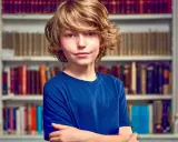 Garçon aux cheveux blonds, bras croisés devant une bibliothèque. Il semble prêt pour un test pour sauter une classe.
