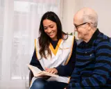 Une jeune femme souriante lit un livre à un homme âgé. Une scène qui illustre comment parler à une fille qu'on ne connaît pas : par le partage et l'écoute.