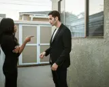 Une femme noire en robe noire pointe du doigt un homme en costume noir, lui reprochant quelque chose. Elle semble vouloir lui faire regretter quelqu'un.