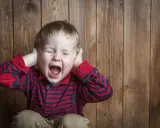 Petit garçon de 5 ans, yeux fermés, criant fort, mains sur les oreilles, en pleine crise de colère incontrôlable.