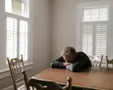 Un homme seul, accablé, le visage enfoui dans ses bras sur une table en bois. La lumière filtre à travers les volets, accentuant la solitude et la méchanceté du PN qui l'a conduit là.