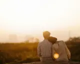 Couple assis dos à dos, la tête de la femme posée sur l'épaule de l'homme, au coucher du soleil.