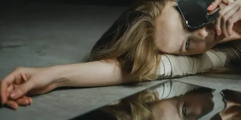 Une femme aux cheveux blonds, le visage tourné, tient un éclat de miroir. Son reflet brisé sur le sol évoque le narcissique et les femmes.