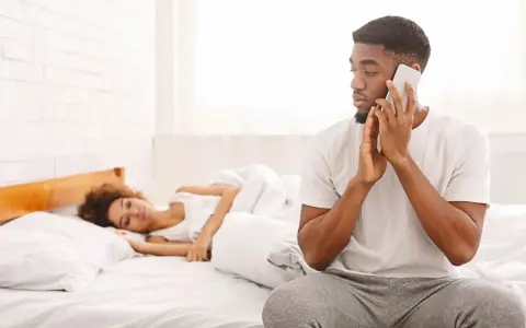 Un homme au téléphone, le regard fuyant, montre un comportement suspect alors qu'une femme dort paisiblement à côté de lui dans le lit.