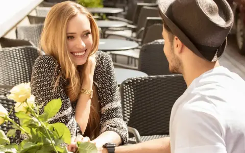 Un couple partage un moment tendre à une terrasse. Elle lui sourit, un bouquet de roses à la main, comme pour lui montrer comment le rendre accro.
