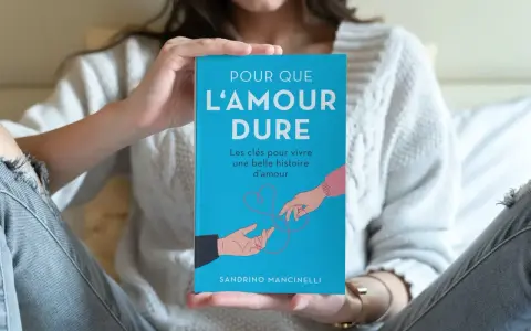Un livre bleu intitulé "Pour que l'amour dure" est tenu par une personne. L'illustration montre deux mains se tendant l'une vers l'autre, symbolisant le début d'une histoire d'amour.