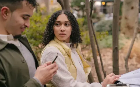 Un homme qui trompe et ment, regardant son téléphone, tandis qu'une femme se méfie, assise à côté de lui dans un parc.