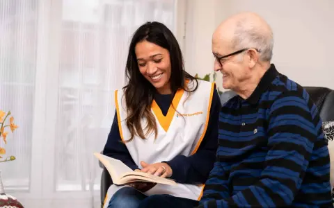 Une jeune femme souriante lit un livre à un homme âgé. Une scène qui illustre comment parler à une fille qu'on ne connaît pas : par le partage et l'écoute.
