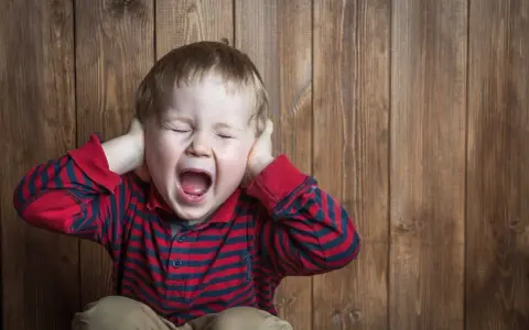 Petit garçon de 5 ans, yeux fermés, criant fort, mains sur les oreilles, en pleine crise de colère incontrôlable.