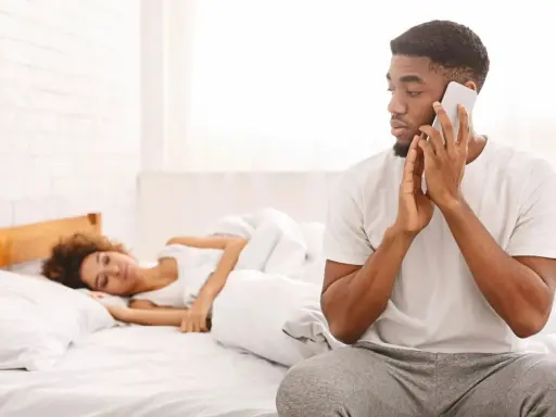 Un homme au téléphone, le regard fuyant, montre un comportement suspect alors qu'une femme dort paisiblement à côté de lui dans le lit.