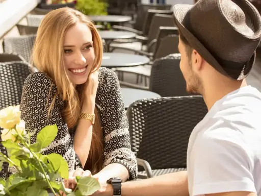 Un couple partage un moment tendre à une terrasse. Elle lui sourit, un bouquet de roses à la main, comme pour lui montrer comment le rendre accro.