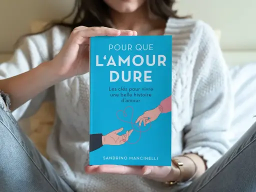 Un livre bleu intitulé "Pour que l'amour dure" est tenu par une personne. L'illustration montre deux mains se tendant l'une vers l'autre, symbolisant le début d'une histoire d'amour.
