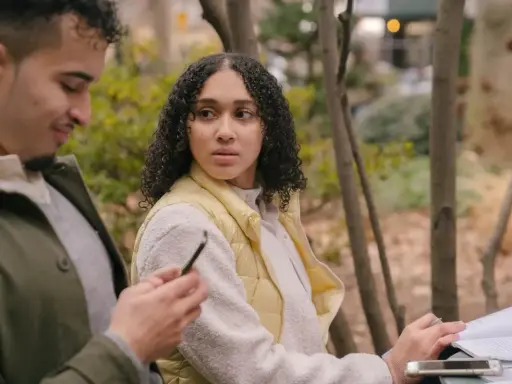 Un homme qui trompe et ment, regardant son téléphone, tandis qu'une femme se méfie, assise à côté de lui dans un parc.
