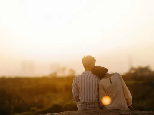 Couple assis dos à dos, la tête de la femme posée sur l'épaule de l'homme, au coucher du soleil.