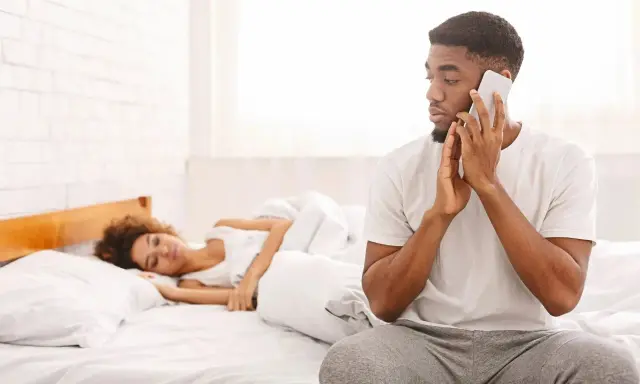 Un homme au téléphone, le regard fuyant, montre un comportement suspect alors qu'une femme dort paisiblement à côté de lui dans le lit.