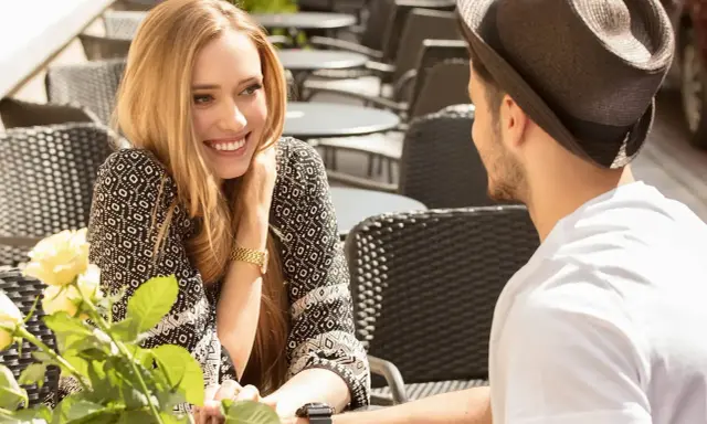 Un couple partage un moment tendre à une terrasse. Elle lui sourit, un bouquet de roses à la main, comme pour lui montrer comment le rendre accro.