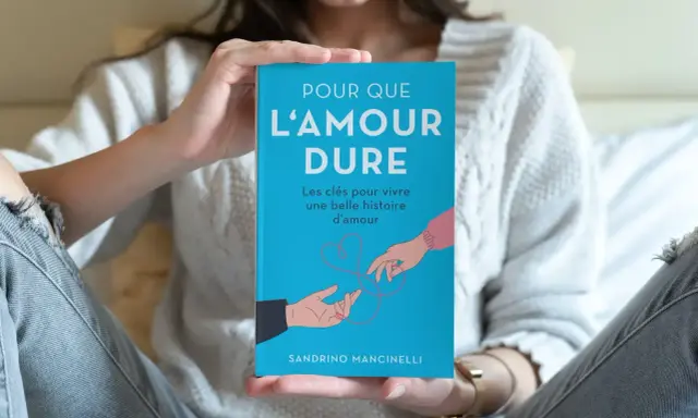 Un livre bleu intitulé "Pour que l'amour dure" est tenu par une personne. L'illustration montre deux mains se tendant l'une vers l'autre, symbolisant le début d'une histoire d'amour.