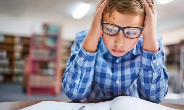 Garçon avec lunettes, mains sur la tête, visiblement en difficulté face à ses devoirs. Signes adolescent HPI, difficultés scolaires.