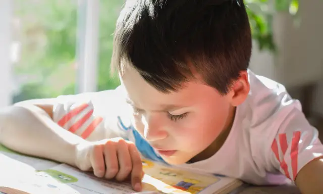 Un garçon concentré lit un livre, son doigt suivant les mots. Un test dys peut être un défi, mais sa détermination est claire.