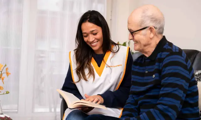 Une jeune femme souriante lit un livre à un homme âgé. Une scène qui illustre comment parler à une fille qu'on ne connaît pas : par le partage et l'écoute.