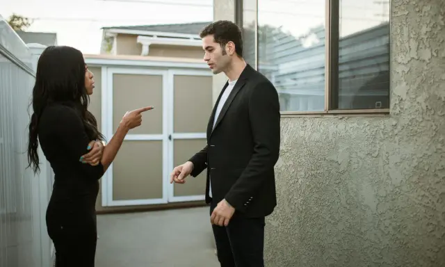 Une femme noire en robe noire pointe du doigt un homme en costume noir, lui reprochant quelque chose. Elle semble vouloir lui faire regretter quelqu'un.