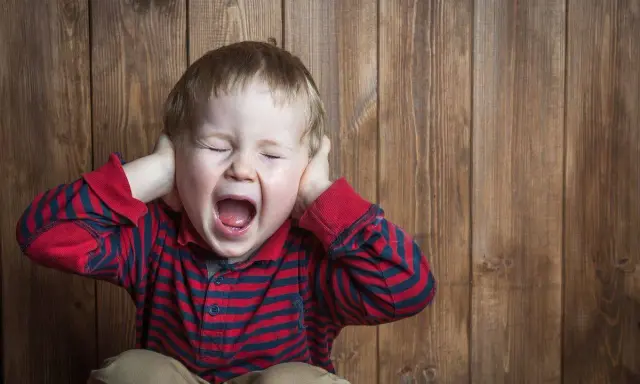 Petit garçon de 5 ans, yeux fermés, criant fort, mains sur les oreilles, en pleine crise de colère incontrôlable.