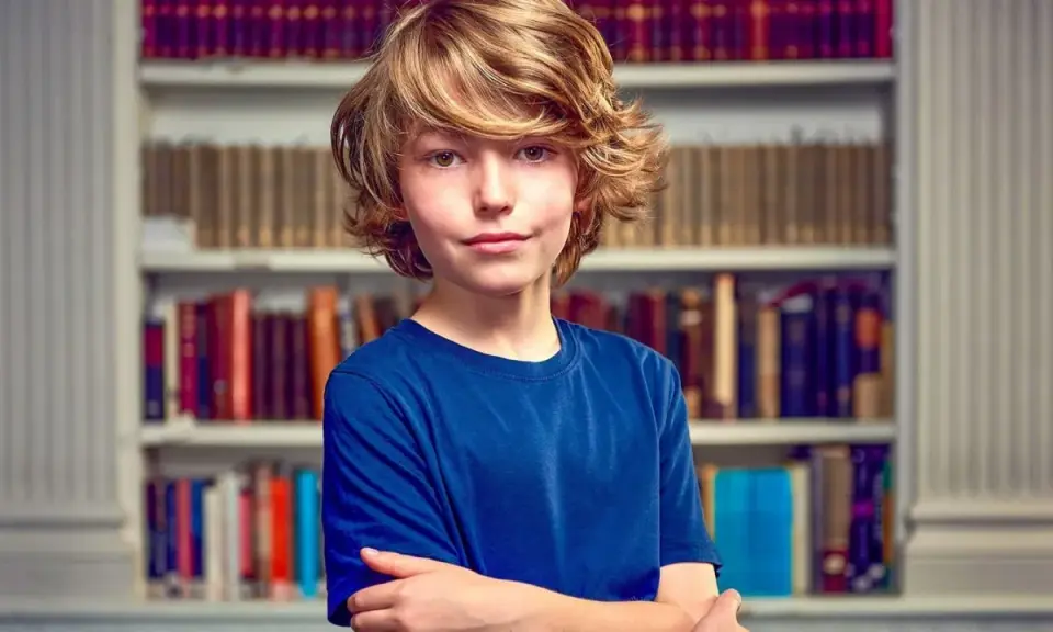 Garçon aux cheveux blonds, bras croisés devant une bibliothèque. Il semble prêt pour un test pour sauter une classe.