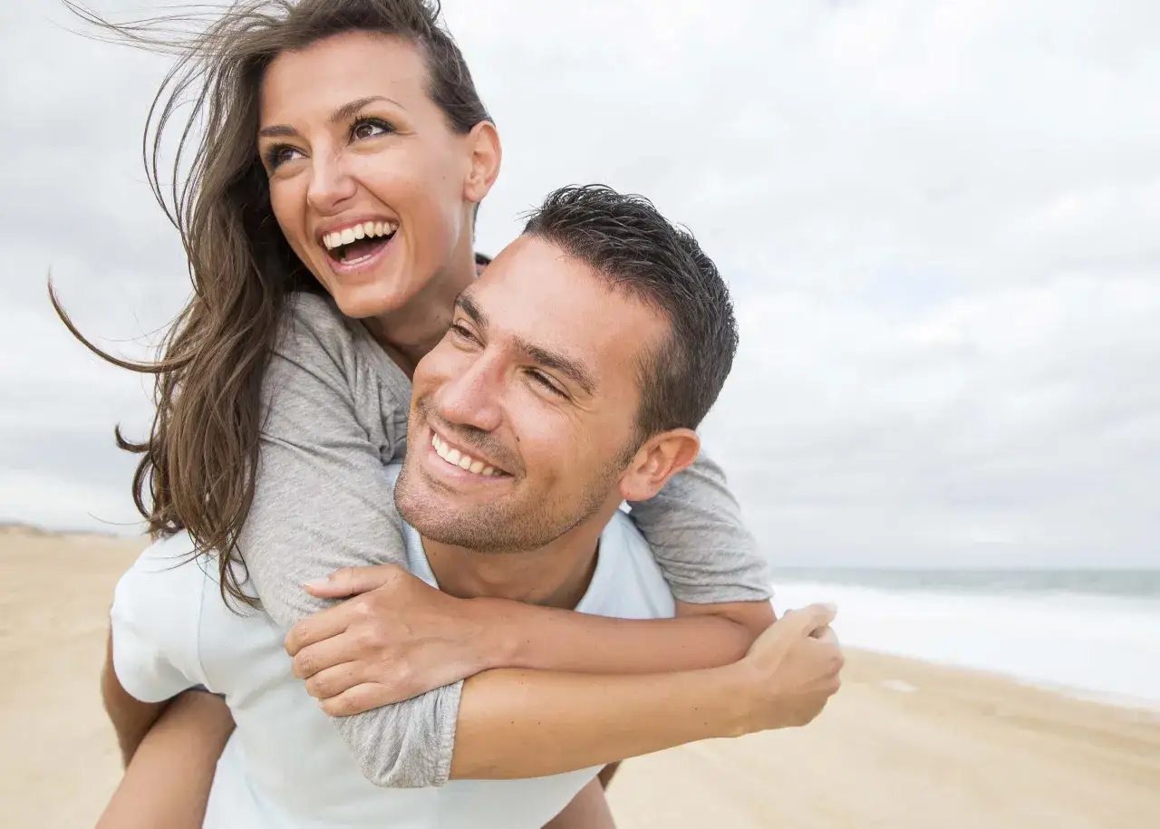 Un couple rit sur la plage, elle est port&eacute;e par lui. Le vent souffle dans ses cheveux. Comment le rendre accro ? Par des moments comme ceux-ci.