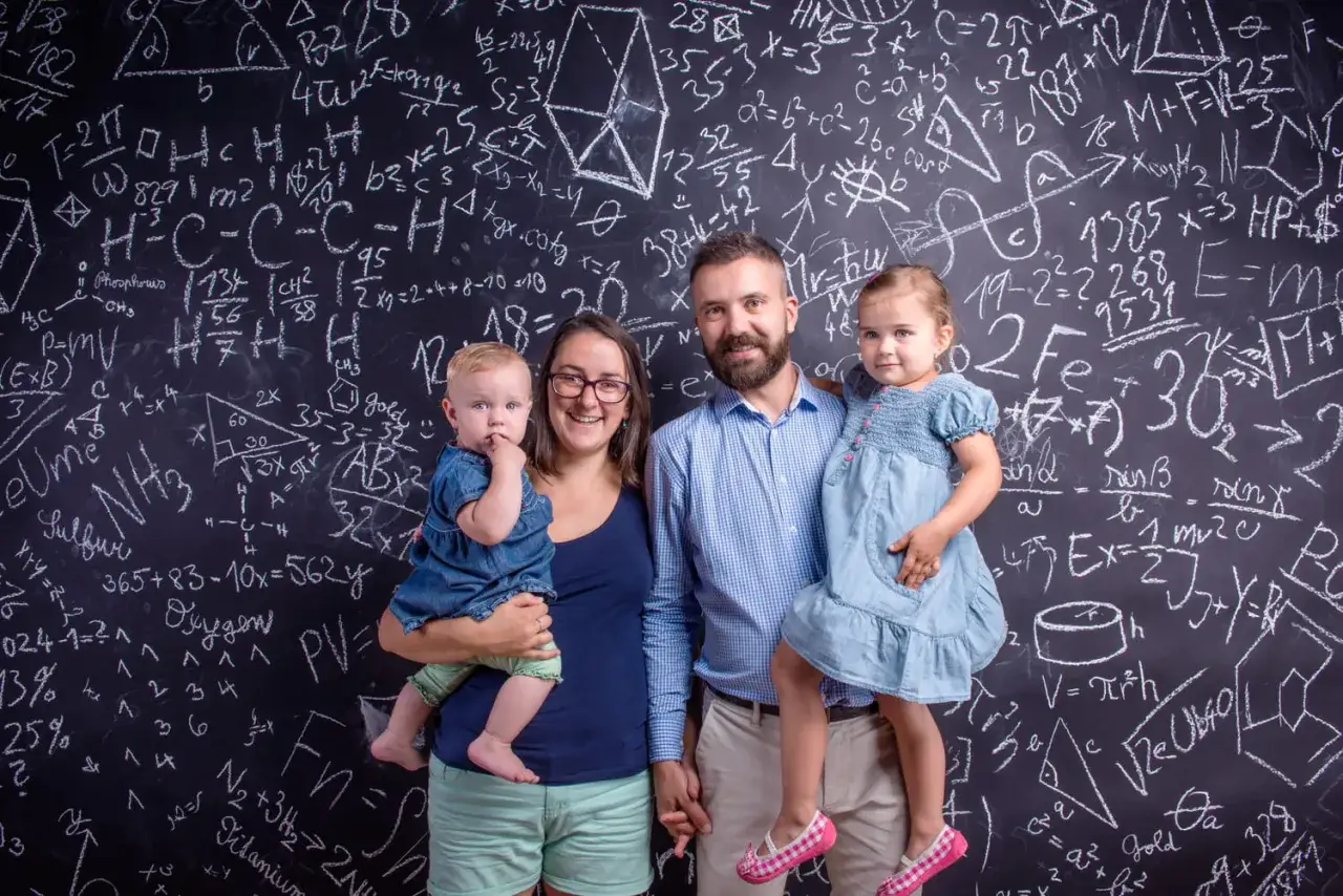 Famille souriante devant un tableau noir rempli d'&eacute;quations. Une m&egrave;re tient un b&eacute;b&eacute;, un p&egrave;re tient une fillette. Une belle illustration de la vie avec un HPI, o&ugrave; la curiosit&eacute; et l'intelligence se c&ocirc;toient.