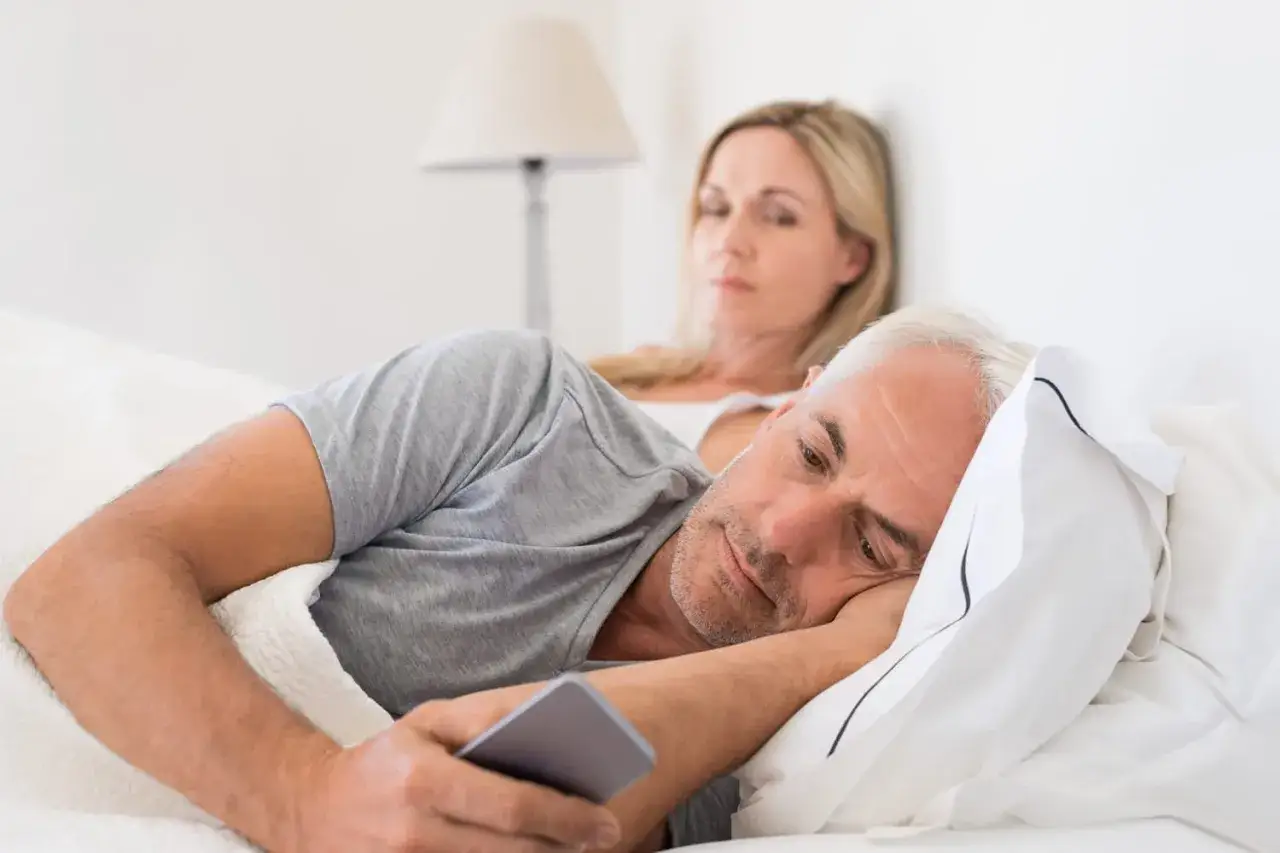 Un homme regarde son t&eacute;l&eacute;phone, l'air pensif, tandis que sa femme le regarde d'un air soup&ccedil;onneux. Des signes d'un homme amoureux de sa ma&icirc;tresse ?