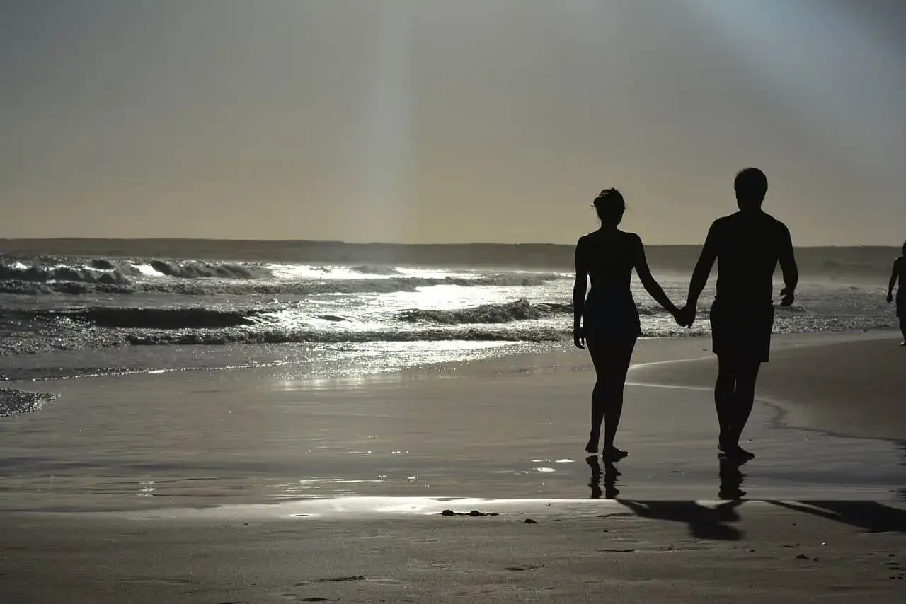 Silhouettes d'un couple main dans la main sur une plage au coucher du soleil. Pourquoi un homme cache qu'il a une copine ? Peut-&ecirc;tre pour pr&eacute;server ce moment de qui&eacute;tude.