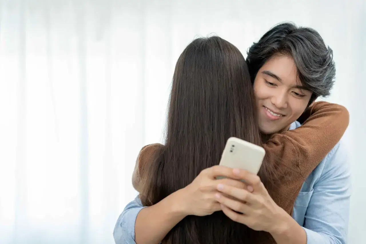Un homme enlace une femme, son regard se porte sur son t&eacute;l&eacute;phone. Comment savoir si mon mari me trompe ? L'image sugg&egrave;re une possible inqui&eacute;tude.