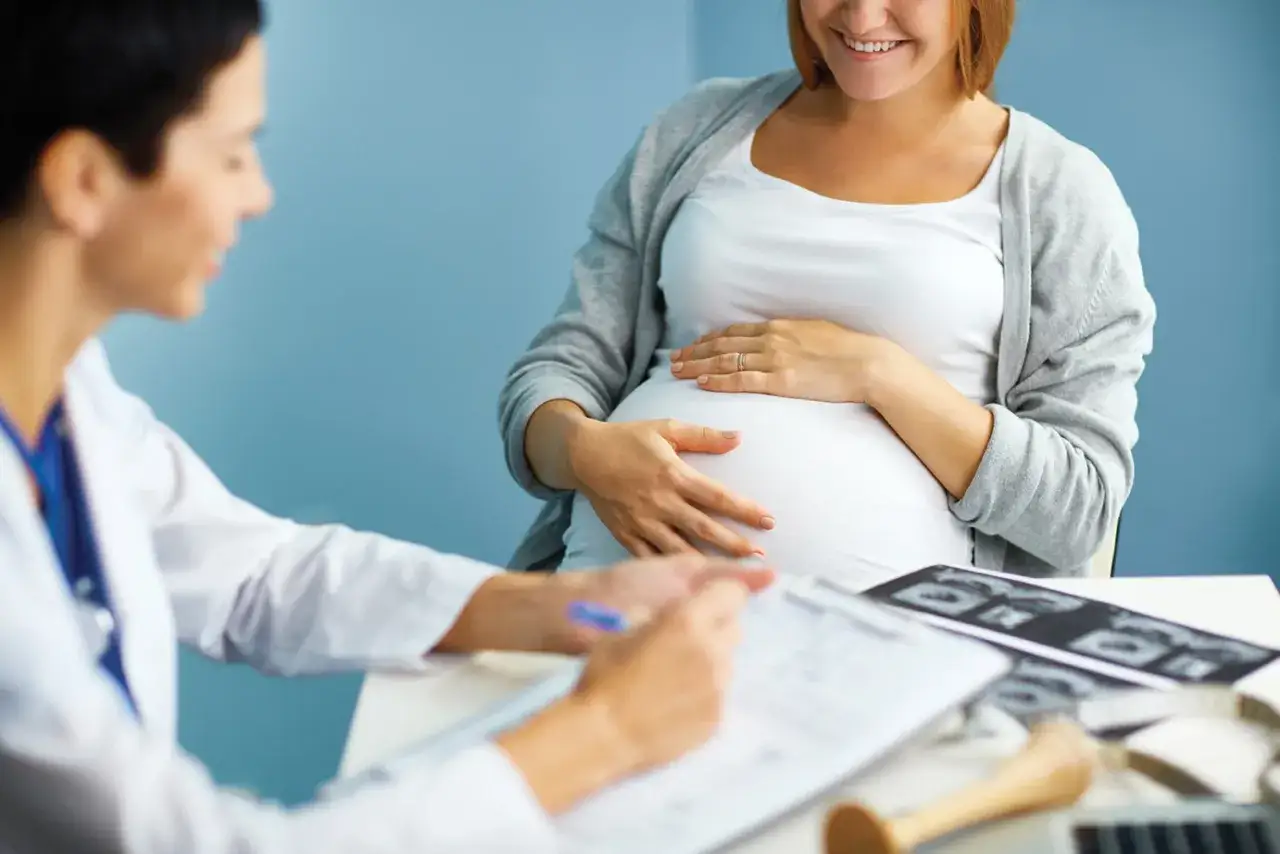 Une femme enceinte sourit en touchant son ventre, lors d'une consultation m&eacute;dicale. Le m&eacute;decin &eacute;crit sur une tablette, un moment cl&eacute; pour apprendre qu'on est enceinte.