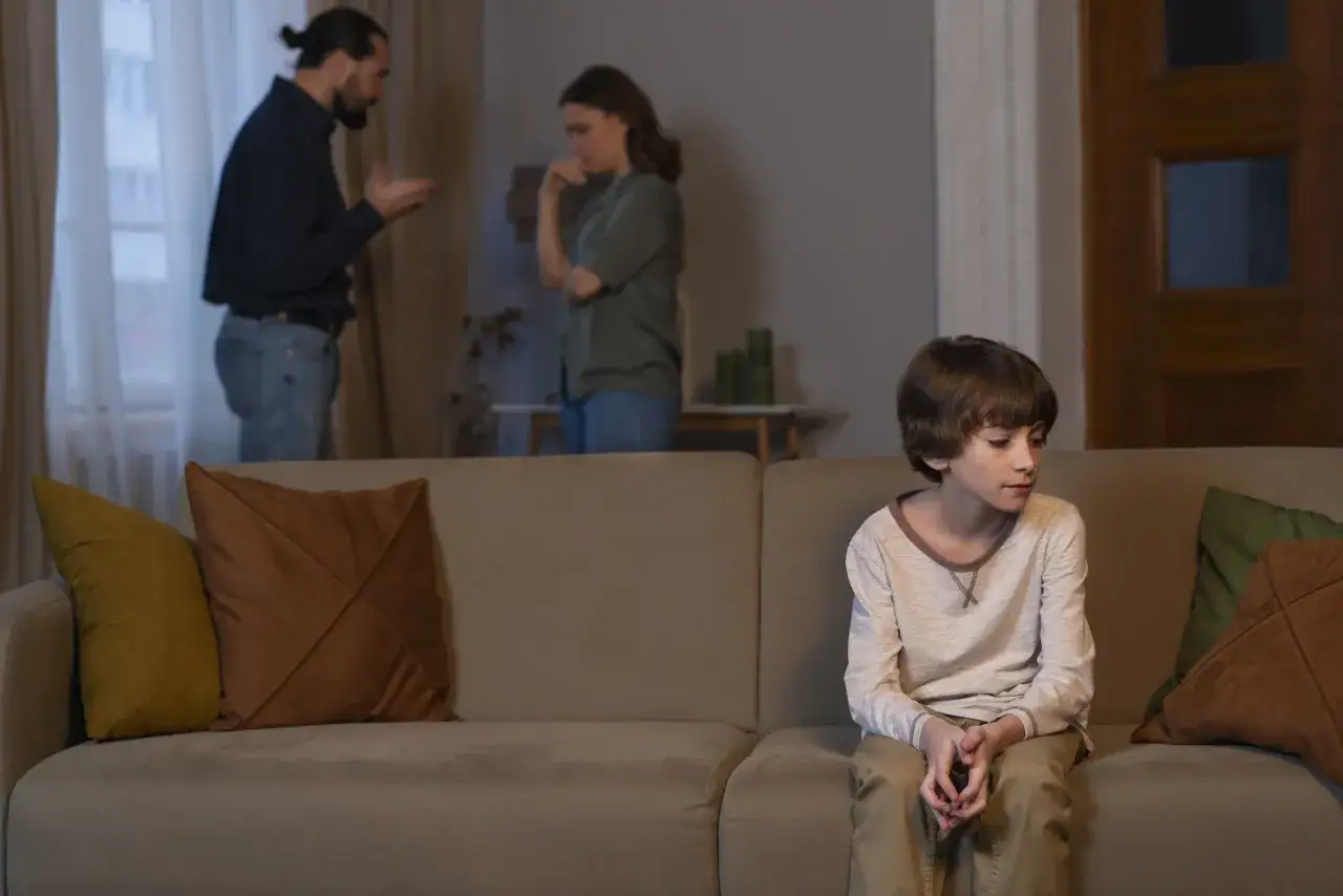 Un enfant seul sur un canap&eacute;, le regard perdu, tandis que ses parents se disputent en arri&egrave;re-plan. Il se sent mal aim&eacute;.