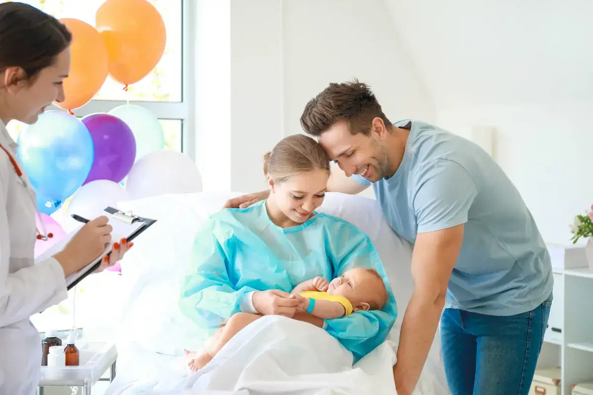 Un couple heureux avec leur nouveau-n&eacute; dans une chambre d'h&ocirc;pital. La maman hospitalis&eacute;e tient le b&eacute;b&eacute;, le papa la regarde tendrement. Des ballons d&eacute;corent la pi&egrave;ce.