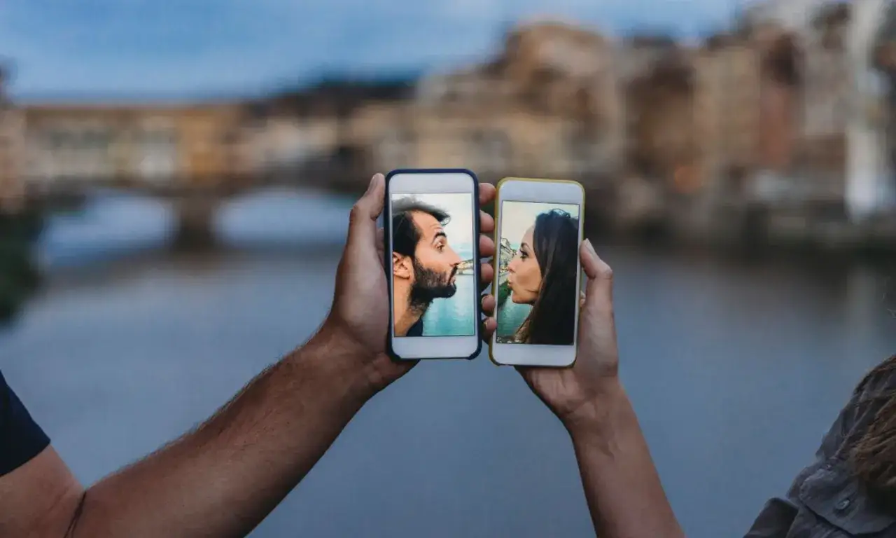 Deux mains tiennent des smartphones affichant des visages se faisant face. Il me d&eacute;bloque sans me parler, cr&eacute;ant un baiser virtuel sur fond de ville.
