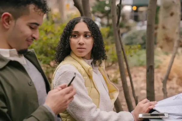 Un homme qui trompe et ment, regardant son téléphone, tandis qu'une femme se méfie, assise à côté de lui dans un parc.